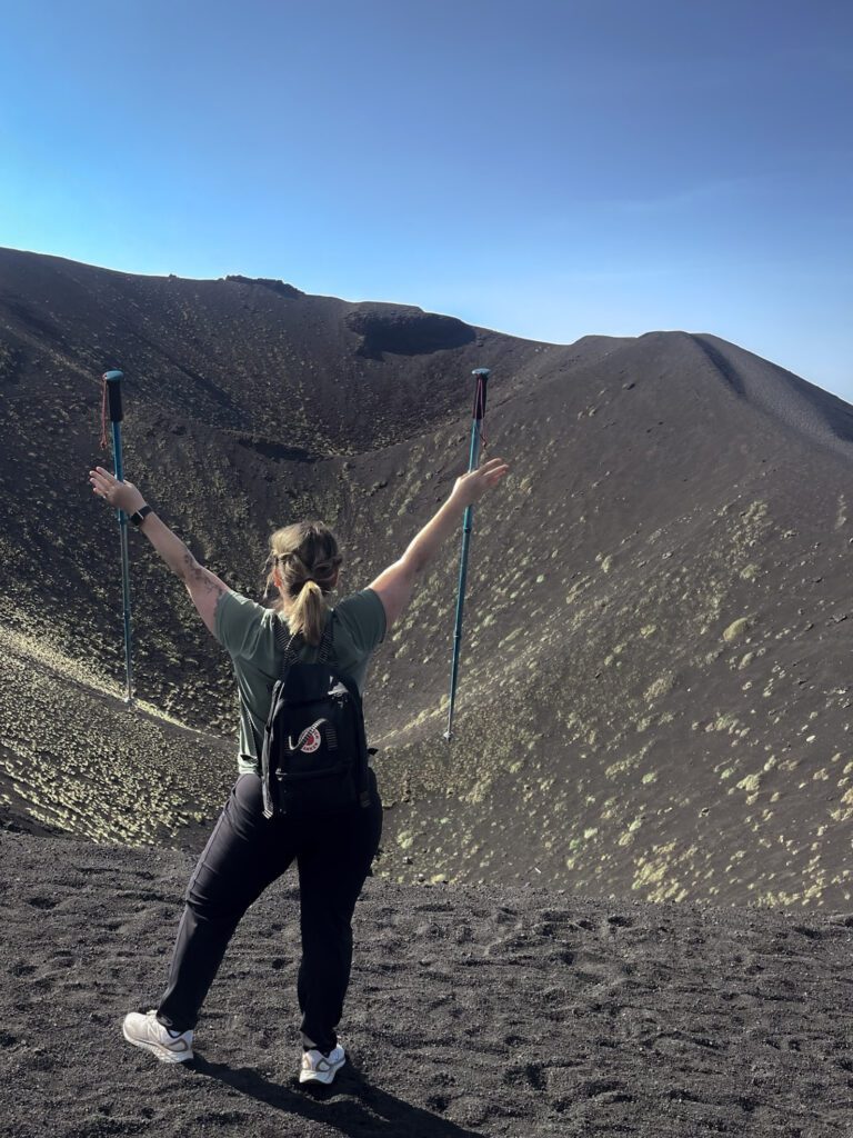 woman with outstretched arms and hiking poles in hand on the black sand of Mt. Etna