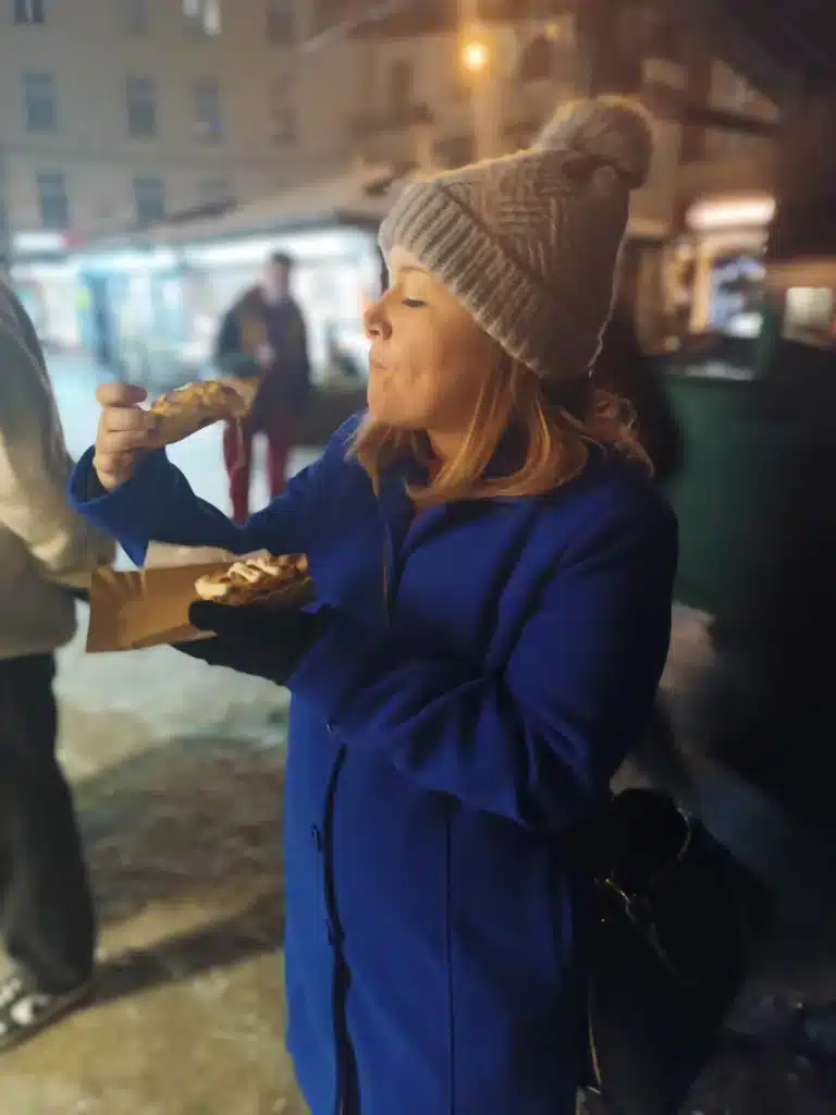 Woman in a bright blue winter coat and cream knit beanie eating a slice of zapiekanka from a paper tray at a food stall in Plac Nowy, Kraków, at night