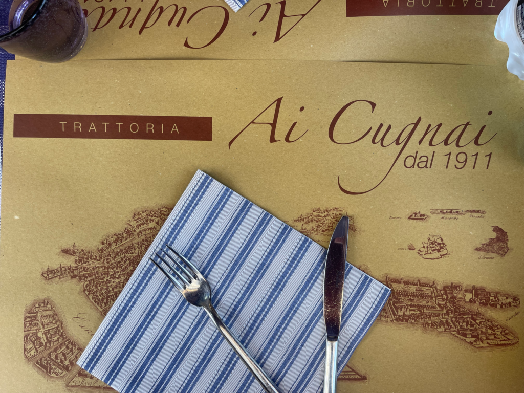 Menu with words Ai Cugnai with a blue and white napkin, and a fork and a knife.