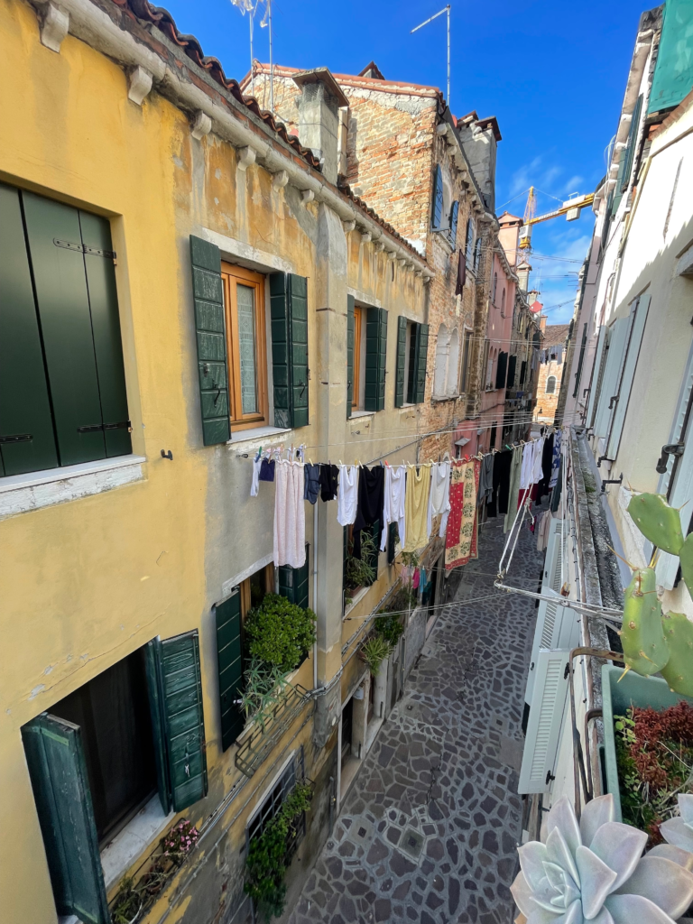 View of a quiet Venetian alley from the apartment with colorful buildings, green shutters, and laundry strung above cobblestone streets, capturing the everyday charm near Arsenale during Carnivale season.