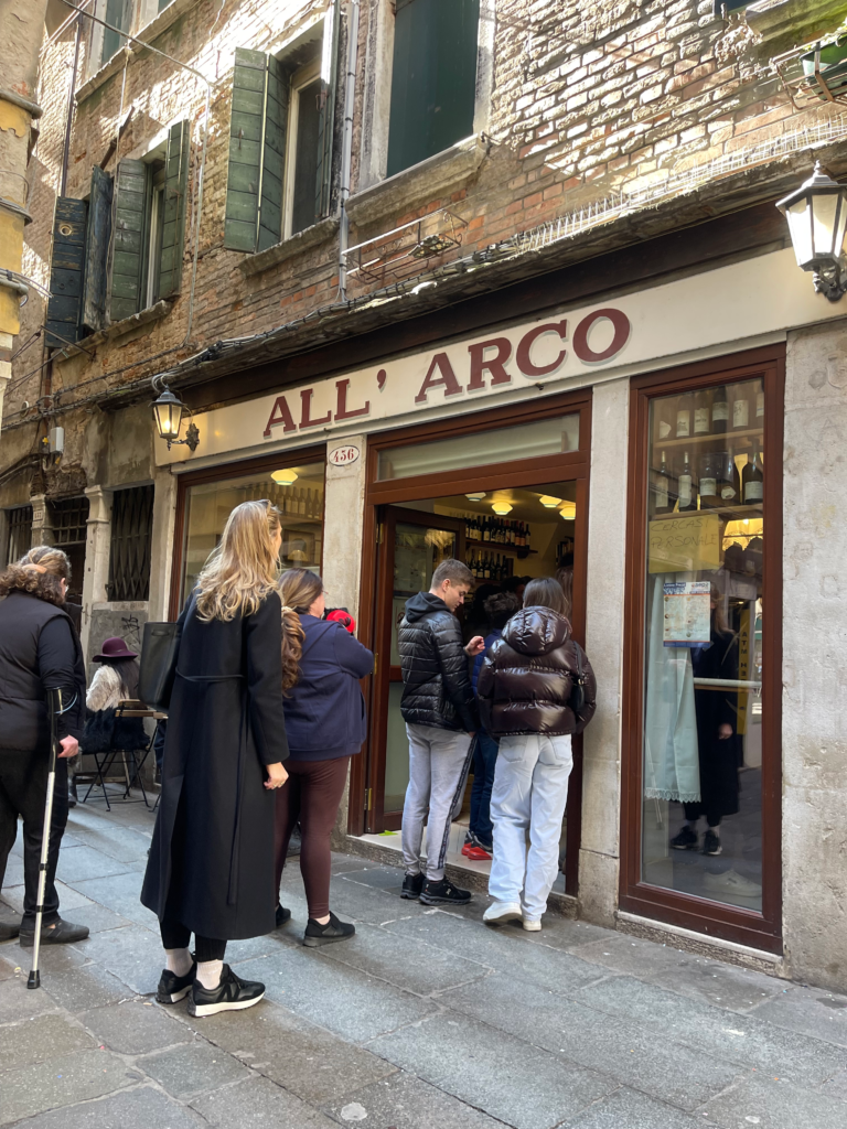 a crowd hanging outside of Bar All' Arco on a Venice Food Tour with a woman patiently waiting to get into the bar dressed in black