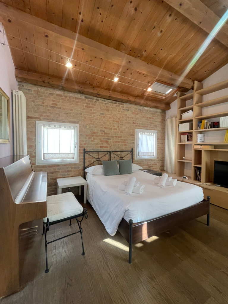Wide view of a bright and rustic bedroom in Fenix Otello, featuring a double bed with white linens, wooden ceiling beams, exposed brick walls, bookshelves, and a piano, ideal for a cozy Venice Carnivale stay.