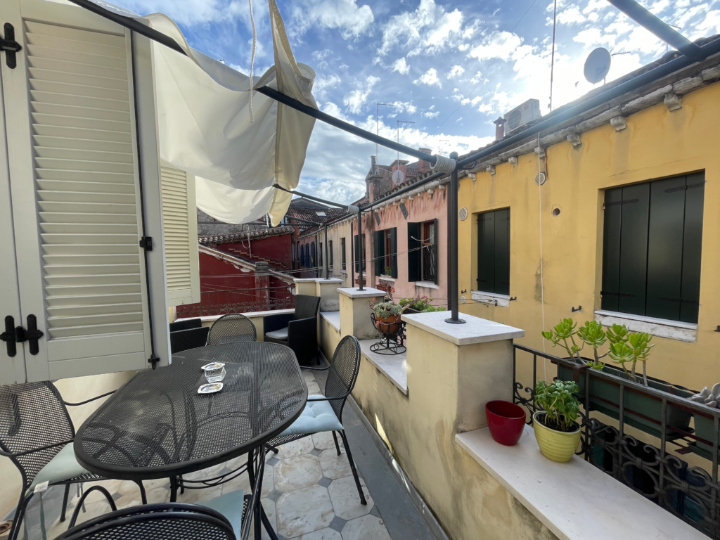Cozy rooftop terrace at Fenix Otello apartment in Venice, featuring a metal dining table with chairs, potted plants, and laundry lines between charming pastel buildings under a bright sky.