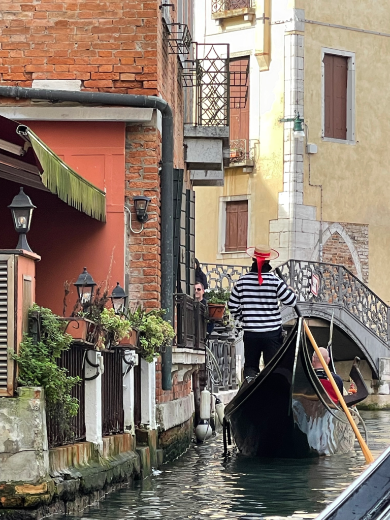 a gondolier near the edge of a sidewalk in a red and white shirt and traditional venetian hat navigating the venetian canals