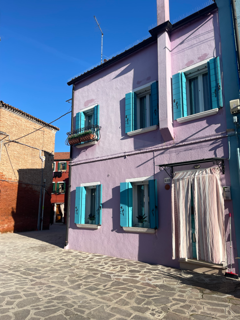 A Purple house in Burano with blue shutters and a white curtain over the door