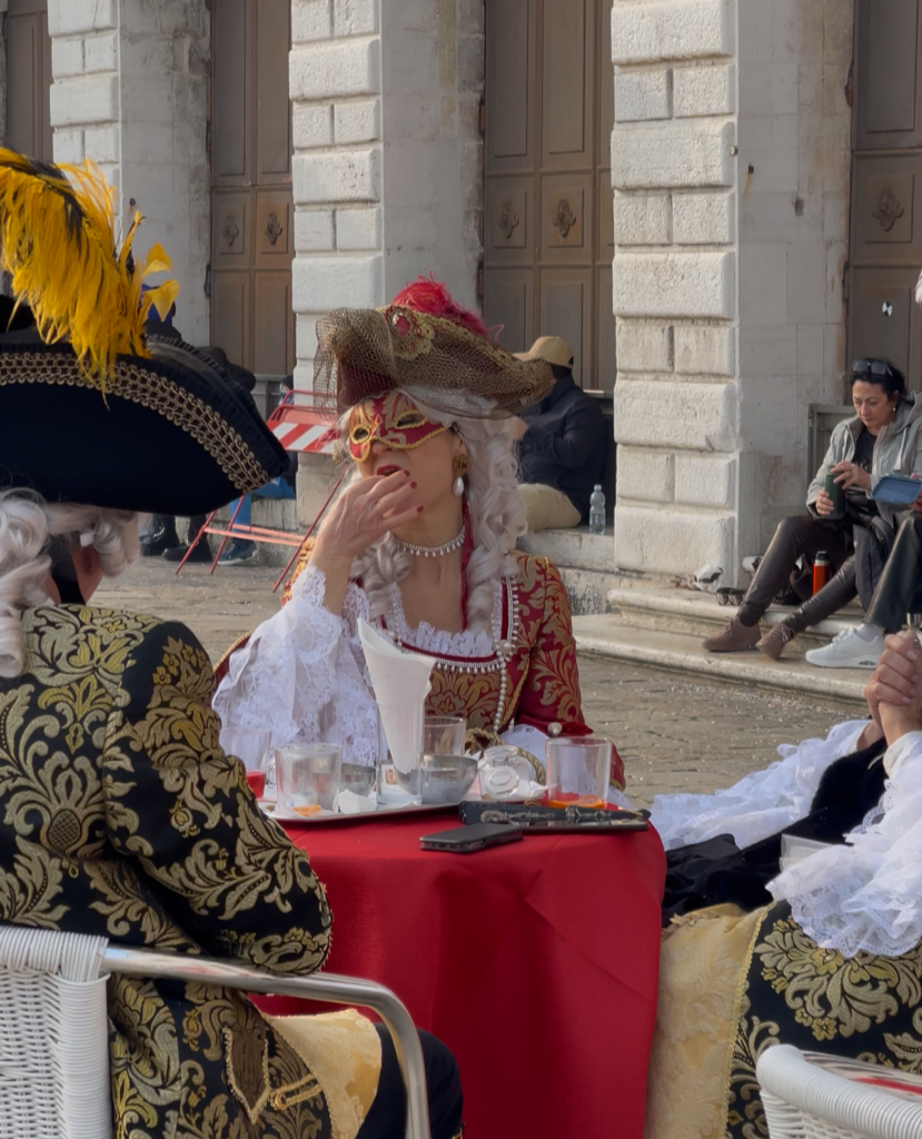 a woman in venetian period costume that is red and white with a veil over her face drinking an aperol in st. marks square in venice italy