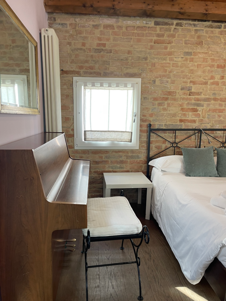 Corner of a bedroom at Fenix Otello with exposed brick walls, a small window with sheer curtains, a wooden upright piano, and a cushioned bench next to a neatly made bed.