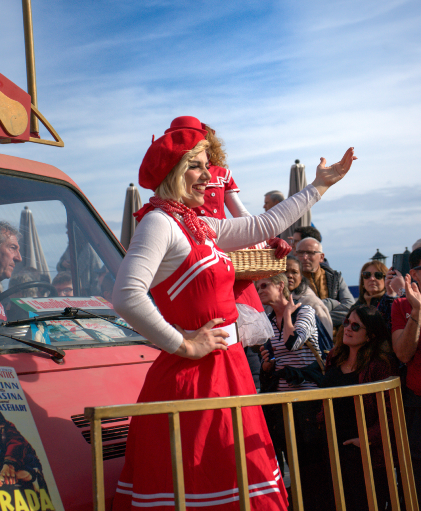 Performer in a vintage red dress and beret gestures to the crowd at the Fête du Citron, standing in front of a retro red van decorated with posters. Spectators watch and smile in the background.