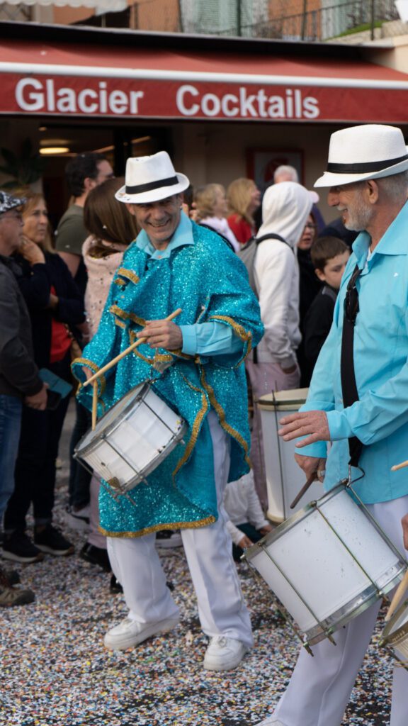 men in blue jackets bang on drums during a parade. They have white hats and white pants. They are smiling