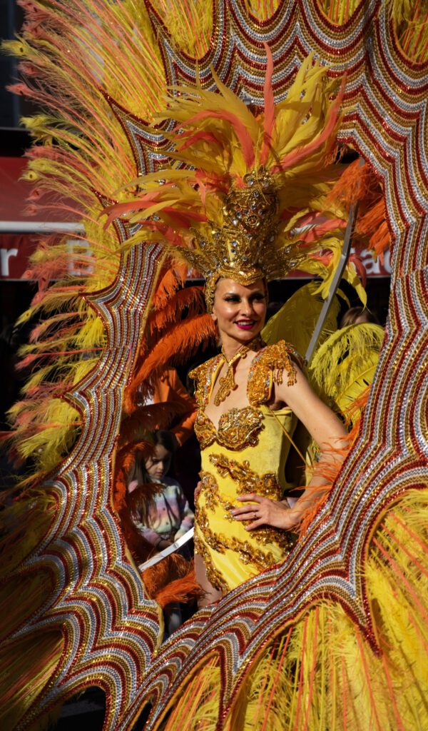 A Young Lady participating in the Fete Du Citron in a vibrant orange costume of feathers and yellow sequins. Smiling at the crowd