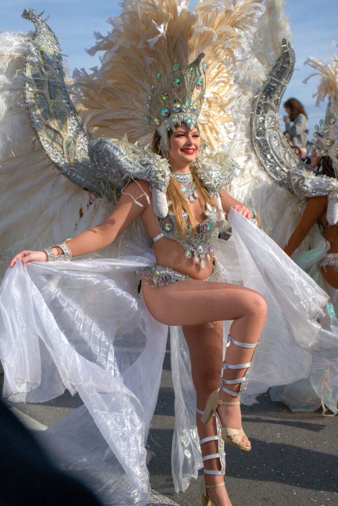 Carnival dancer in a silver and white feathered costume smiles as she poses with one leg raised during the Fête du Citron parade. Her outfit sparkles with sequins and gems under the sun.