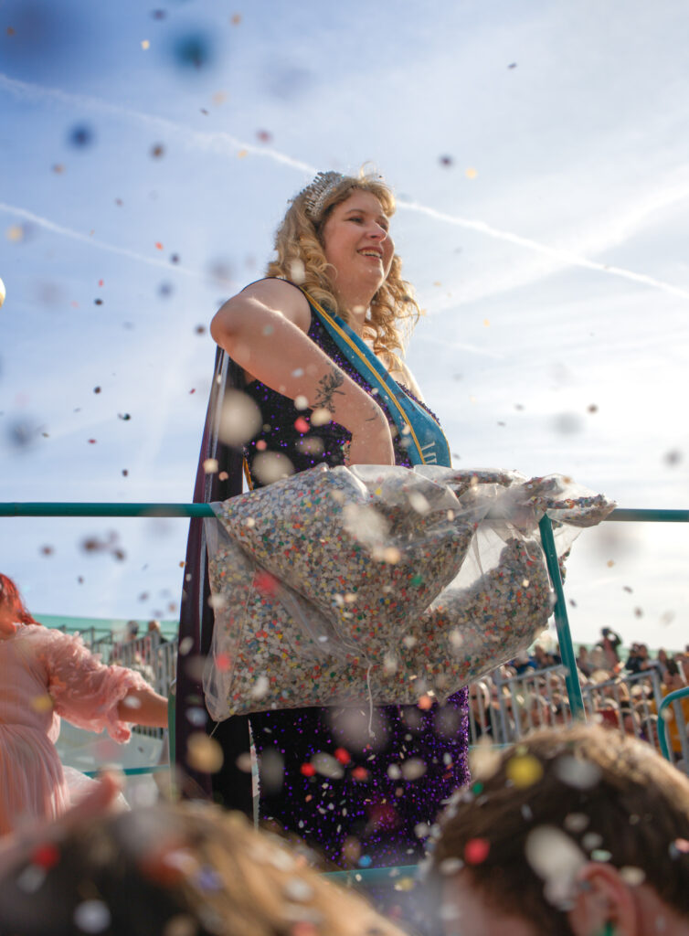 Woman wearing a sparkling tiara and sash smiles from a parade float at the Fête du Citron, surrounded by a shower of colorful confetti. She holds a large plastic bag filled with more confetti as the crowd watches below.