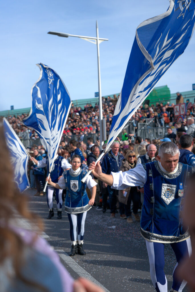 Flag bearers in blue and white uniforms march in formation at the Fête du Citron parade, waving large blue banners with white designs. Spectators fill the stands along the street.