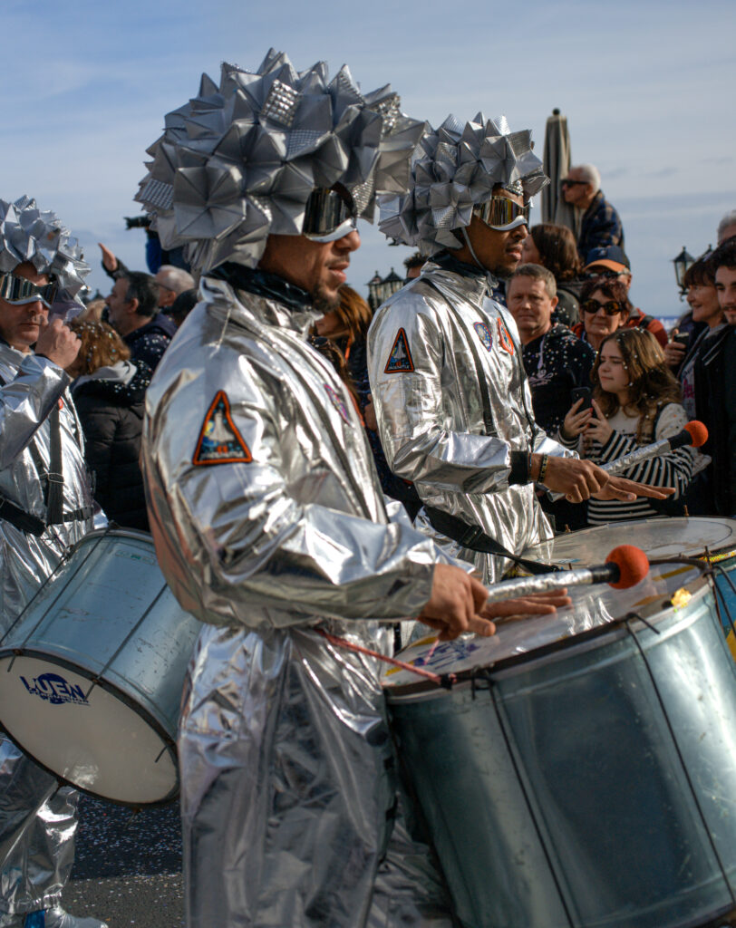 Drummers dressed in futuristic silver costumes with geometric helmets and mirrored visors perform in the Fête du Citron parade. They march past a crowd while playing metallic drums with orange-tipped mallets.