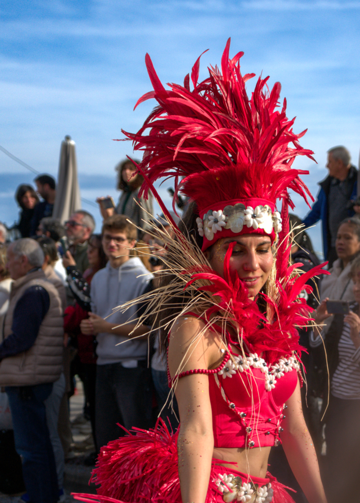 Performer in a vibrant red costume adorned with feathers, seashells, and straw details smiles during the Fête du Citron parade. Her headpiece and outfit draw attention as she walks past a crowd of onlookers.