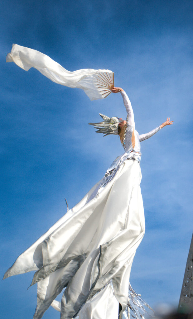 Performer on stilts dressed in an ethereal white and silver costume raises a fan with flowing fabric against a clear blue sky at the Fête du Citron. Her dramatic metallic headdress and sweeping garments create a striking, celestial look.