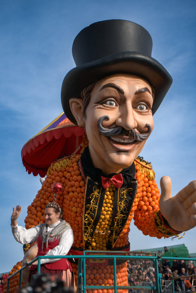 A large parade float at the Fête du Citron features a towering figure with a top hat made entirely of oranges, accompanied by a woman in traditional dress waving from below.