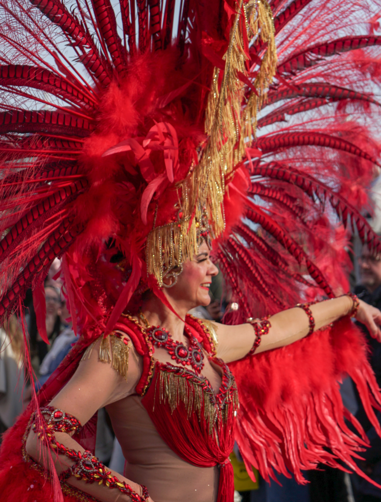 Smiling woman in a red feathered costume with golden accents stands in profile during the Fête du Citron parade. Onlookers behind her watch the performance with interest.