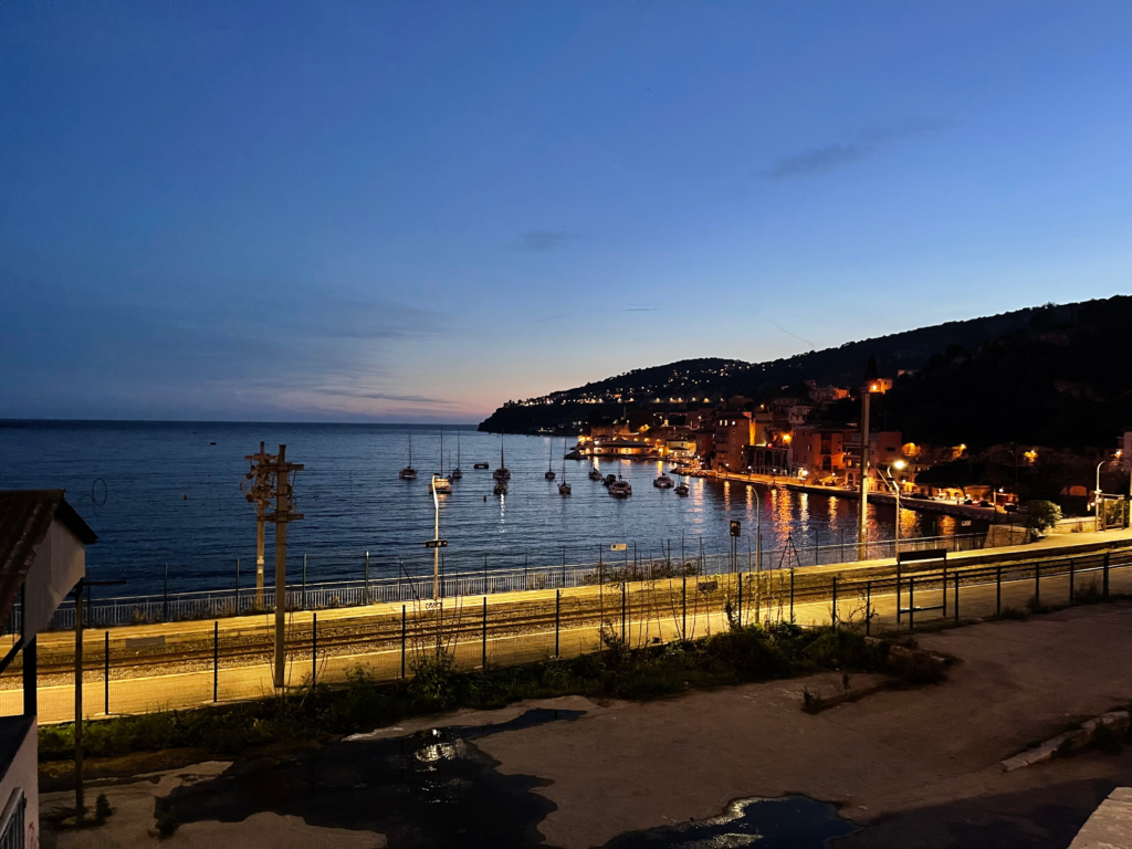 View of Villefranche-sur-Merโs harbor at twilight, with sailboats floating in the calm bay and the waterfront glowing with warm lights, seen from above the coastal train tracks.