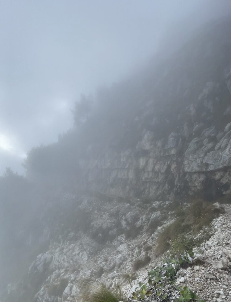 A rocky mountain path, reminiscent of Italys Road of 52 Tunnels, is enveloped in thick fog, obscuring the view. The terrain is rugged, with patches of greenery visible among the rocks. The atmosphere is misty and mysterious, with limited visibility.