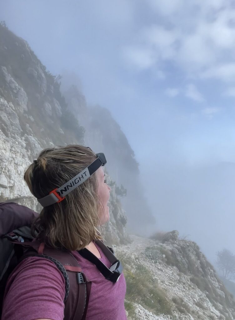 A person wearing a headlamp and backpack stands on a rocky, foggy trail along the Road of 52 Tunnels, gazing at the misty landscape. The sky is partially visible with patches of blue and clouds.