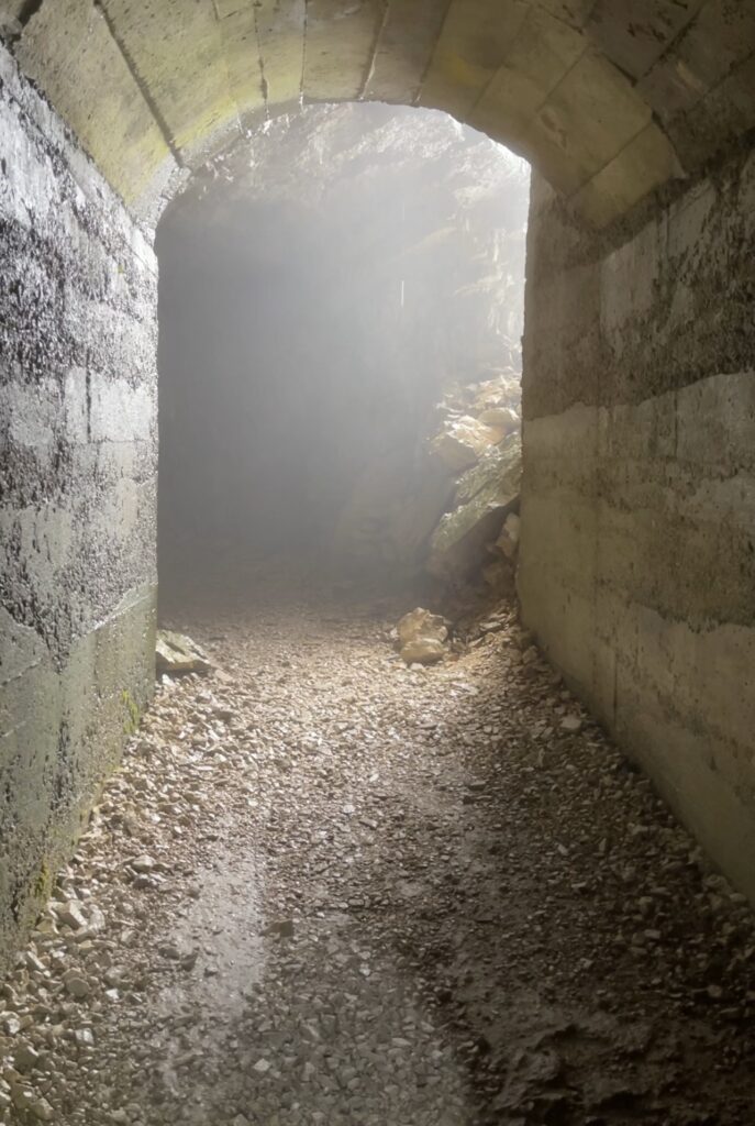 A dimly lit tunnel with rough stone walls leads to an exit, where natural light and mist create a hazy effect. The ground is covered with gravel and small rocks, reminiscent of the trails on the Strada Delle 52 Galleries in Italy. The archway at the end opens to a rocky, misty scene.