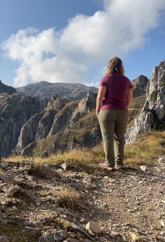 A person with long hair stands on a rocky mountain path, wearing a purple shirt and green pants. Facing away, they gaze at the rugged mountains under a partly cloudy sky. The scene along Strada Delle 52 Galleries conveys tranquility and the beauty of hiking in Italys nature.