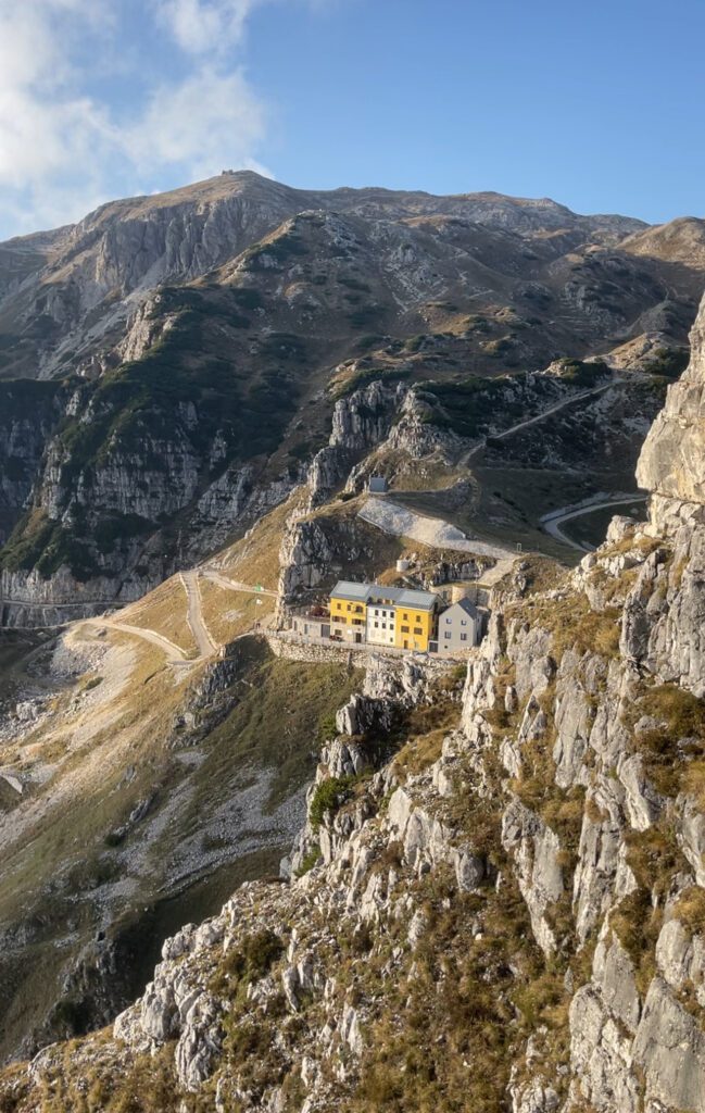 Mountain landscape with a steep rocky slope and winding paths. Two adjacent buildings, one painted yellow and the other white, are nestled in a valley. Beyond the charming Rifugio Achille Papa, rugged mountain peaks rise under a blue sky, perfect for hiking in Italy.