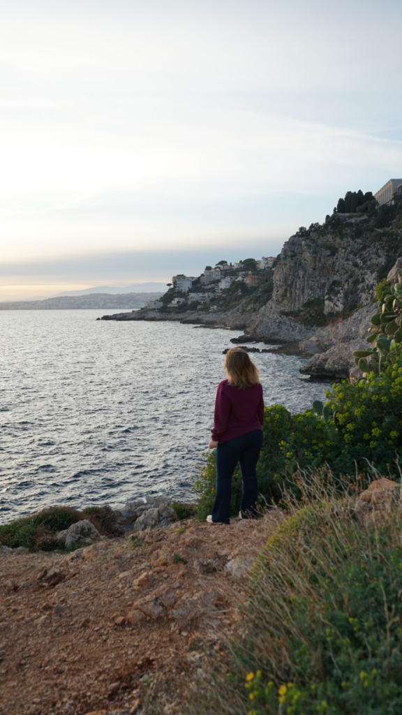 Person in a burgundy hoodie stands on a rocky cliff along the Sentier du Littoral, gazing over the Mediterranean Sea toward the rugged coastline of Villefranche-sur-Mer at dusk.