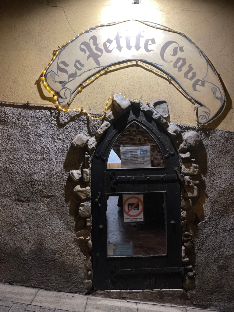 Stone-framed doorway under a curved sign reading “La Petite Cave,” softly lit by fairy lights against a textured stucco wall. A no credit card sign is posted on the dark wooden door, marking the entrance to the restaurant.
