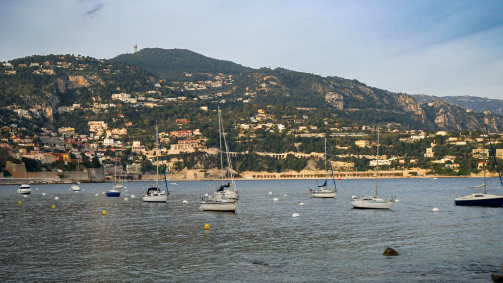 Sailboats gently anchored in the calm bay of Villefranche-sur-Mer, with terraced hillside homes and rugged cliffs in the background.