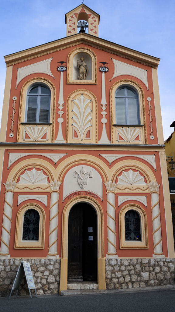 The faรงade of Chapelle Saint-Pierre in Villefranche-sur-Mer, featuring coral and cream colors, artistic detailing, and a small statue above the doorway.
