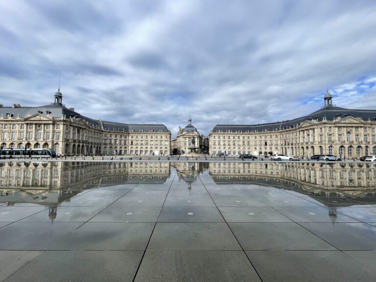 The image displays the Place de la Bourse in Bordeaux, France, featuring its classical architecture with intricate facades. In the center stands a prominent building with a dome and a statue on top. The square is mirrored perfectly on the water surface in front, creating a symmetrical reflection under a cloudy sky. This scene is interesting for its blend of historical architecture and modern reflective installation, showcasing an iconic city landmark known for its beauty and cultural significance.