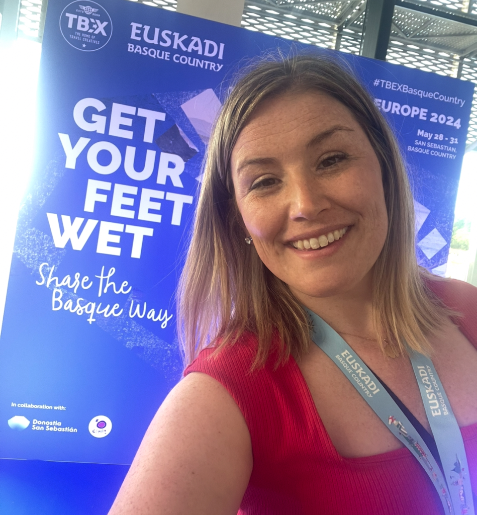 woman smiling with a red shirt on with a background that says " GET YOUR FEET WET, EXPLORE THE BASQUE WAY"