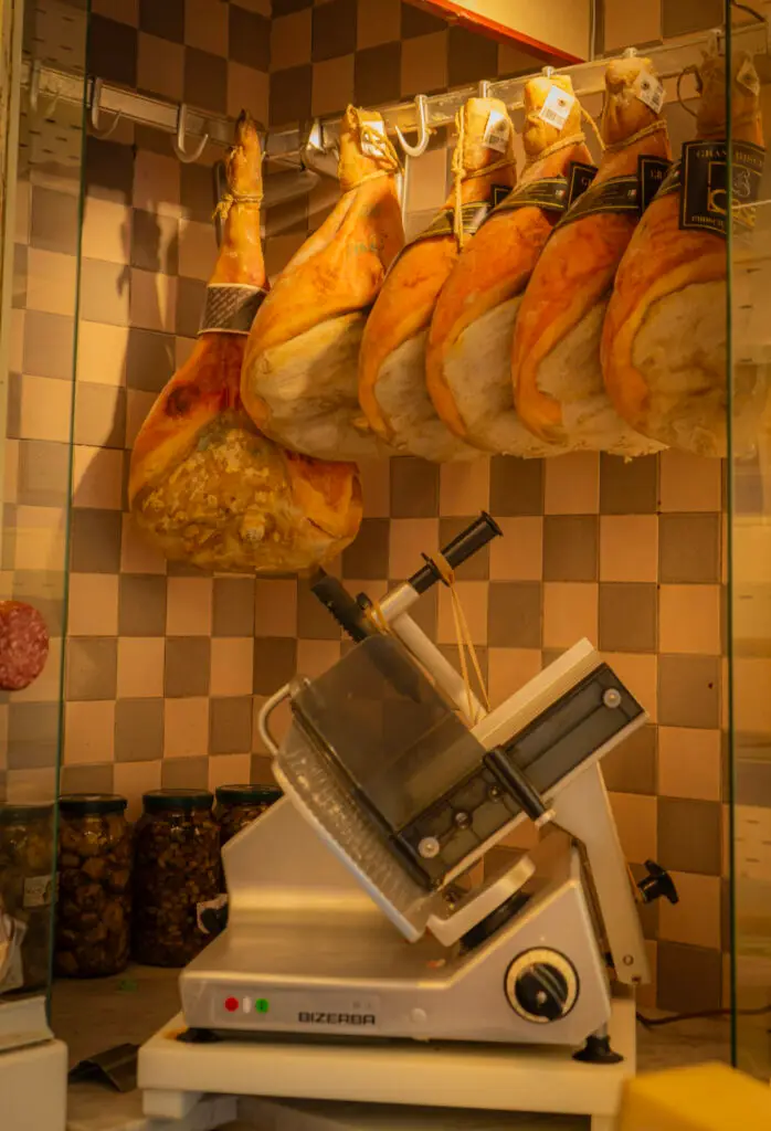 Inside an Italian cheese shop, a slicer in the corner with hanging meats during one of the best tours in venice