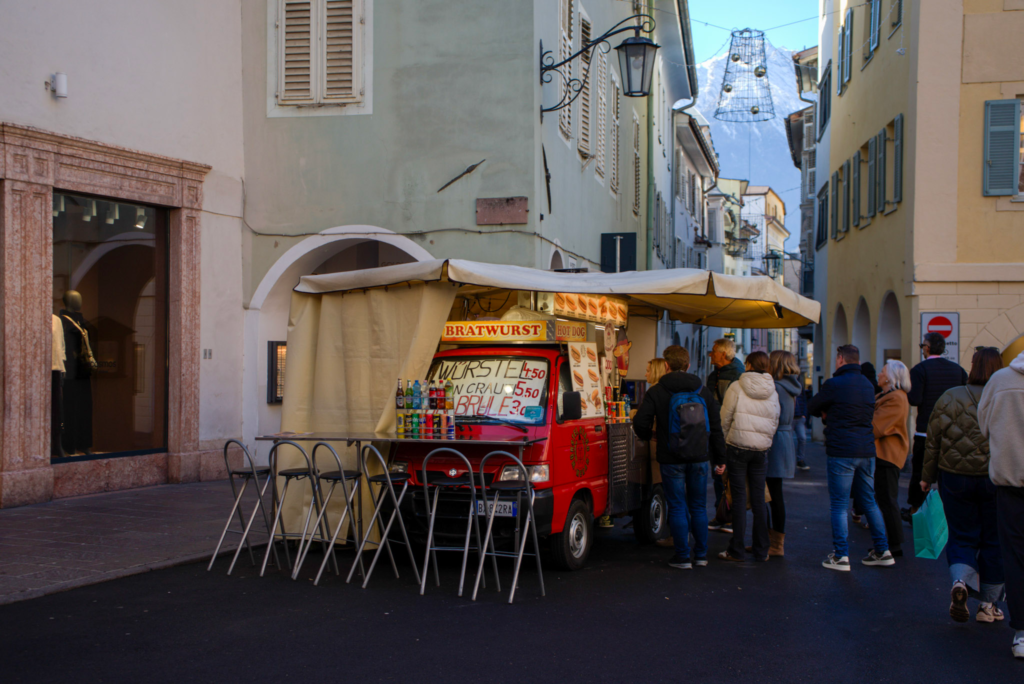 A small red food truck with a “Bratwurst” sign and handwritten prices for sausages, sauerkraut, and drinks serves a line of people on a narrow street in Merano during the Christmas market. The truck is surrounded by folding chairs and bottles of soda, with festive decorations hanging above the street and snow-capped mountains visible in the background.