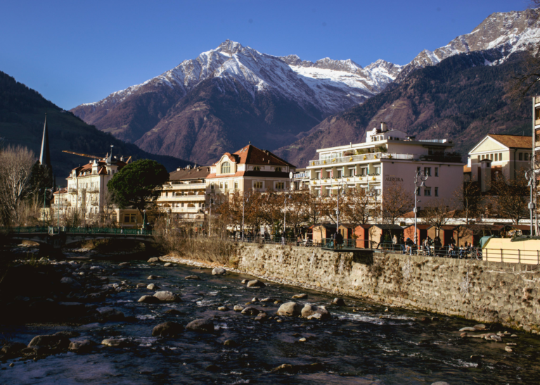 Scenic view of Merano, Italy, with the Passer River in the foreground and snow-capped Alps in the background. Elegant buildings line the riverbank, including Hotel Aurora, near the site of the Merano Christmas Market.