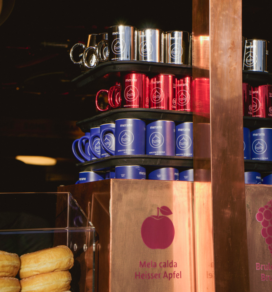 Stacked souvenir mugs in silver, red, and blue on display at the Merano Christmas Market, each labeled with “Merano” and “Mercatino di Natale.” Below the mugs, a copper stand features an apple graphic and bilingual text reading “Mela calda” and “Heisser Apfel,” indicating hot apple drinks.