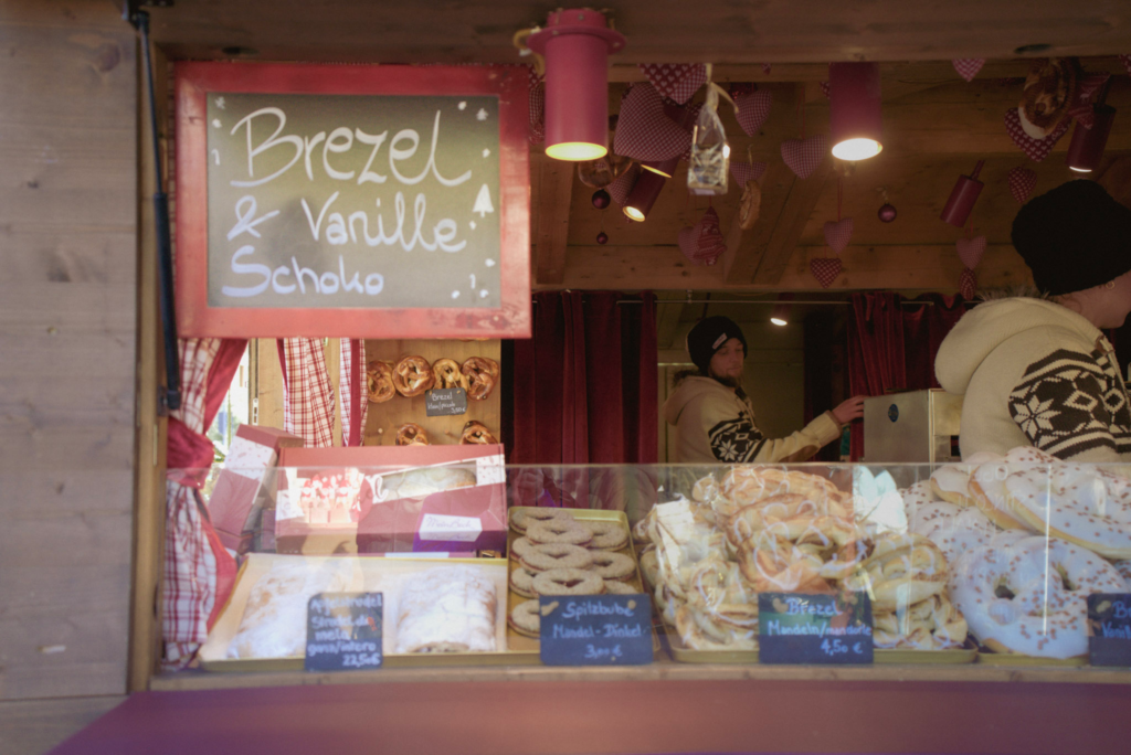 Bakery stall at the Merano Christmas Market selling a variety of festive treats including chocolate, vanilla, and almond pretzels, Spitzbuben cookies, and apple strudel. A chalkboard sign reads “Brezel & Vanille Schoko,” while two vendors in winter sweaters work inside a wooden booth decorated with red gingham hearts.