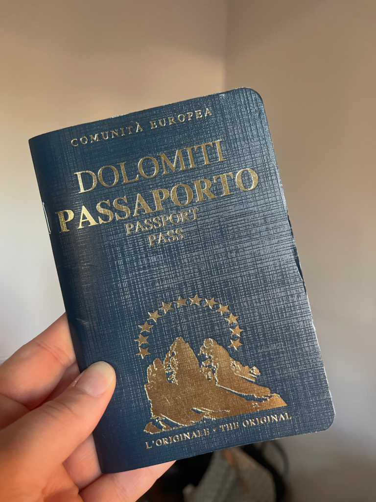 A close-up of the Dolomiti Passaporto, a deep blue booklet with gold-embossed text and an illustration of mountain peaks under a circle of stars. The passport is held by a hand and resembles a European-style travel document.