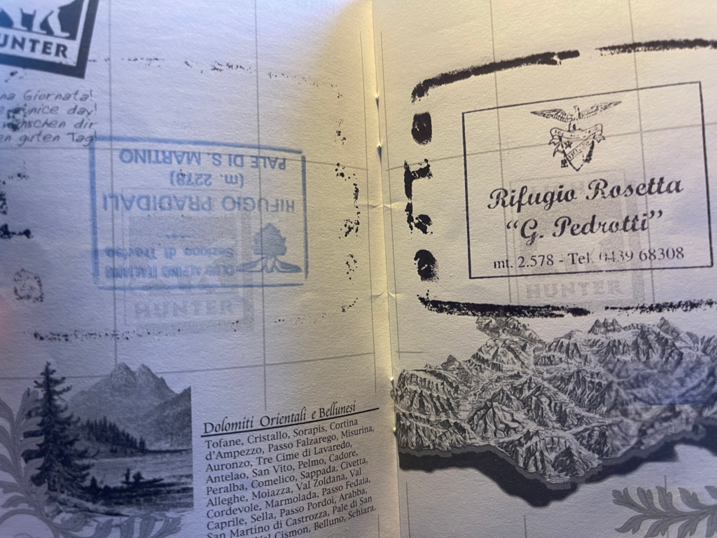 Open pages of the Dolomiti Passaporto showing stamps from Rifugio Pradidali and Rifugio Rosetta "G. Pedrotti," along with sketched illustrations of mountain ranges and a list of Dolomite locations like Tofane and Cristallo.