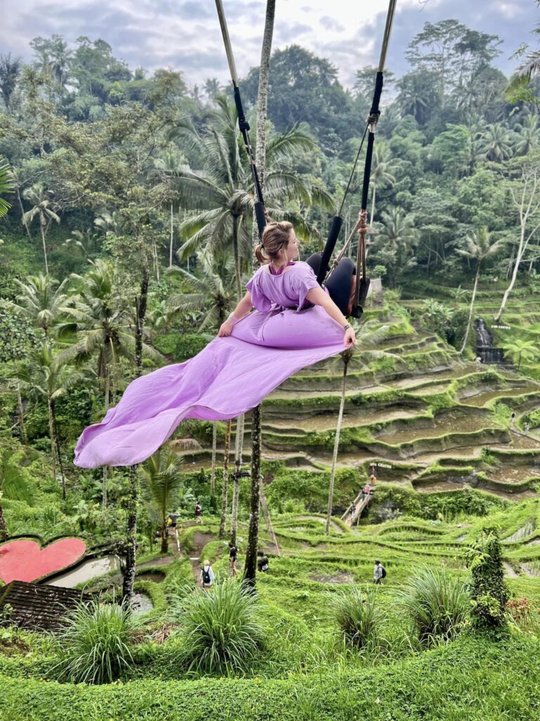 Author swings through the terraced rice fields of Ubud in Bali. She's wearing a Pink dress that is flowing behind her.