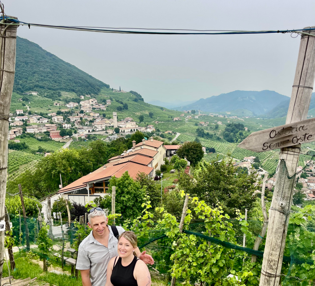 A man in a light shirt and a woman with blonde hair and wearing all black stand in front of rows of vineyards at Senz D' Osteria in Valdobbiadene