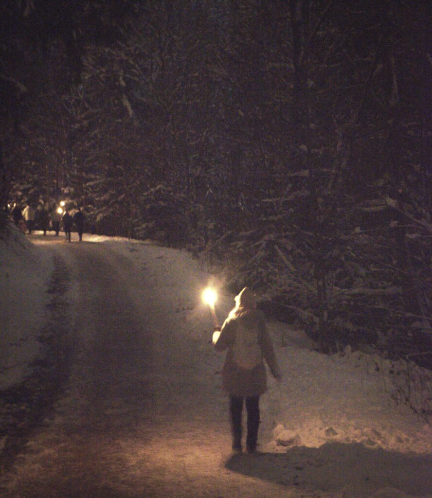 A young woman holds a torch, illuminating the snow  as she walks towards the group of particpants holding torches on their way to the Ravenna Gorge