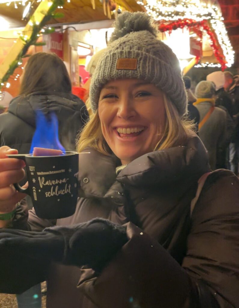 a woman holding a cup of mulled wine that is on fire. She is smiling and has a grey cap on with a black jacket