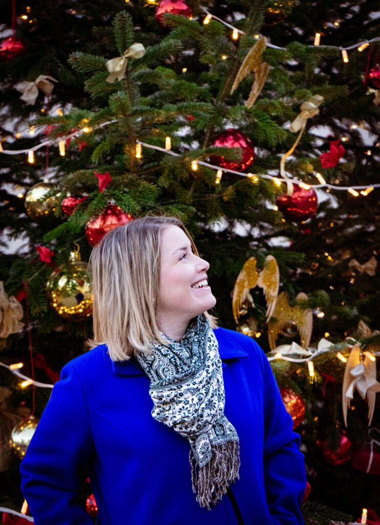 Woman with blonde hair in blue jacket standing in front of a christmas tree looking off into the distance and she has a green scarf on. 