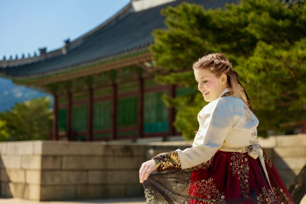 Woman in red and black hanbok standing next to a palace, holding her dress. 