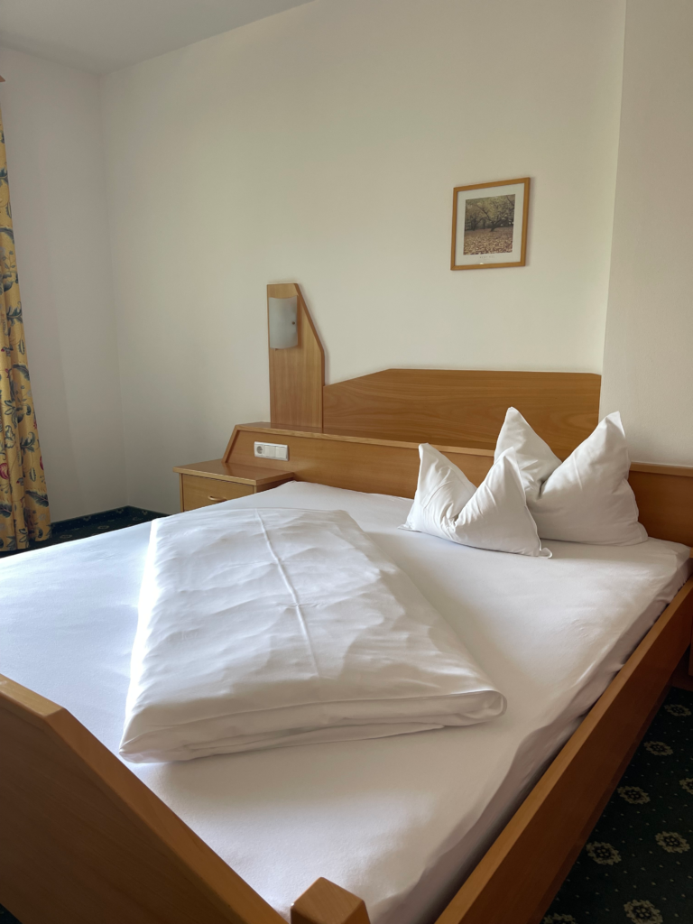 Cozy wooden hotel room at Hotel Trenker with crisp white bedding, two large pillows, and a neatly folded duvet on the bed. Light wood furniture and soft natural light create a warm and simple base before starting Alta Via 1 near Lago di Braies.