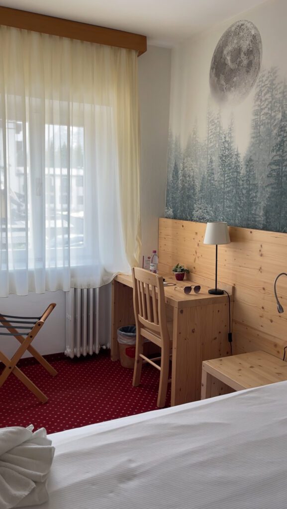 Light grain desk with a white curtain in the background with a bed that has white sheets on it.