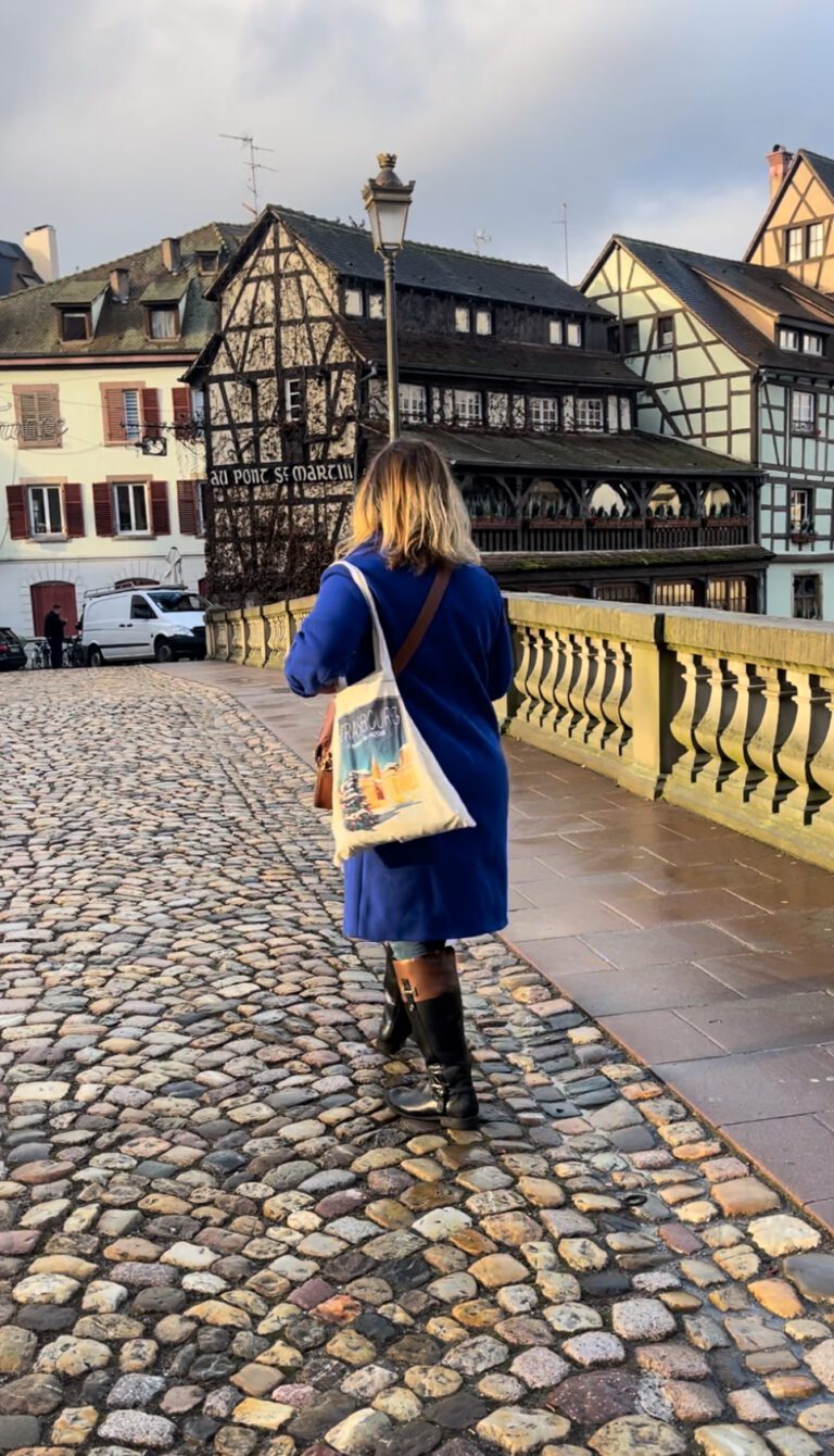 Woman walking across a cobblestone street in a blue coat with a tote that says Strasbourg at a Christmas Market